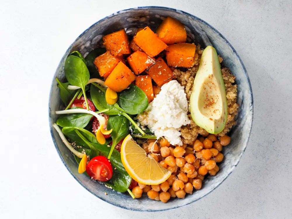 A vibrant organic sweet potato and chickpea bowl with quinoa, spinach, avocado, and cherry tomatoes, topped with a lemon wedge.