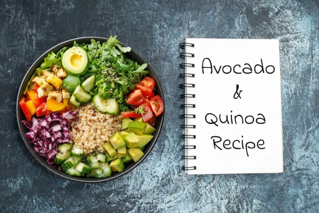 Overhead view of a fresh avocado and quinoa salad bowl packed with cherry tomatoes, cucumber, and mixed greens, placed next to a recipe notebook.