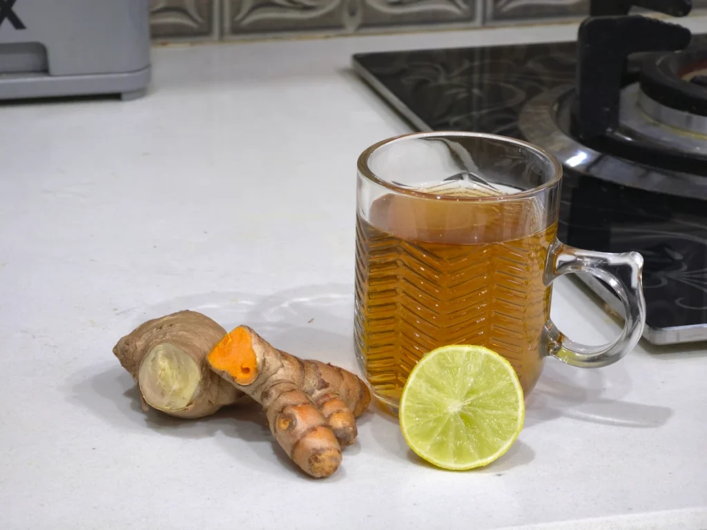 A freshly brewed cup of homemade turmeric ginger tea sitting on a white kitchen counter next to a stovetop, with raw ginger, turmeric pieces, and a slice of lemon.