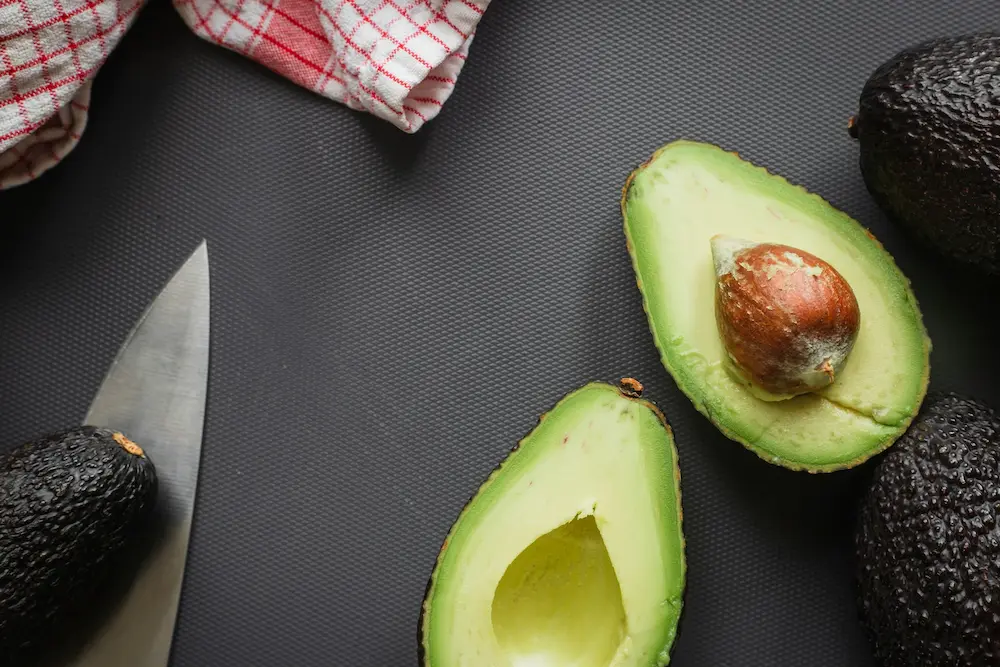 A top-down close-up of freshly halved avocados resting on a dark textured surface next to a chef's knife, highlighting their rich, heart-healthy monounsaturated fats.