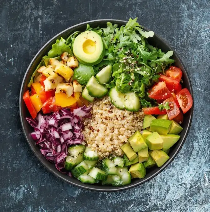Close-up overhead view of a freshly prepared avocado and quinoa salad recipe in a dark bowl, featuring diced cucumbers, cherry tomatoes, and mixed greens.