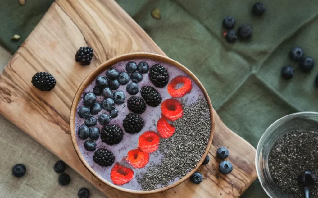 Overhead view of a thick organic mixed berry smoothie bowl in a wooden bowl, carefully topped with distinct rows of fresh raspberries, blackberries, blueberries, and raw chia seeds, resting on a wooden cutting board.