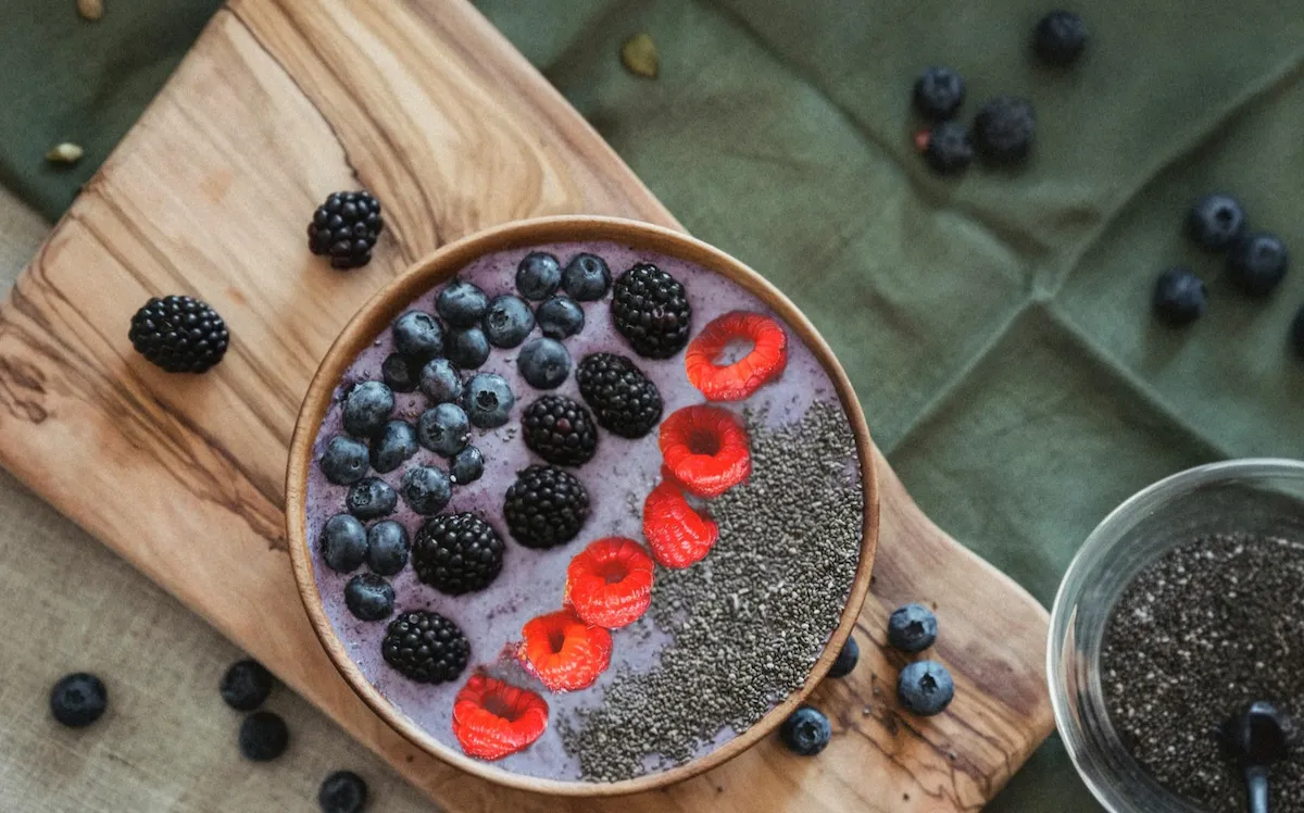 Overhead view of a thick organic mixed berry smoothie bowl in a wooden bowl, carefully topped with distinct rows of fresh raspberries, blackberries, blueberries, and raw chia seeds, resting on a wooden cutting board.