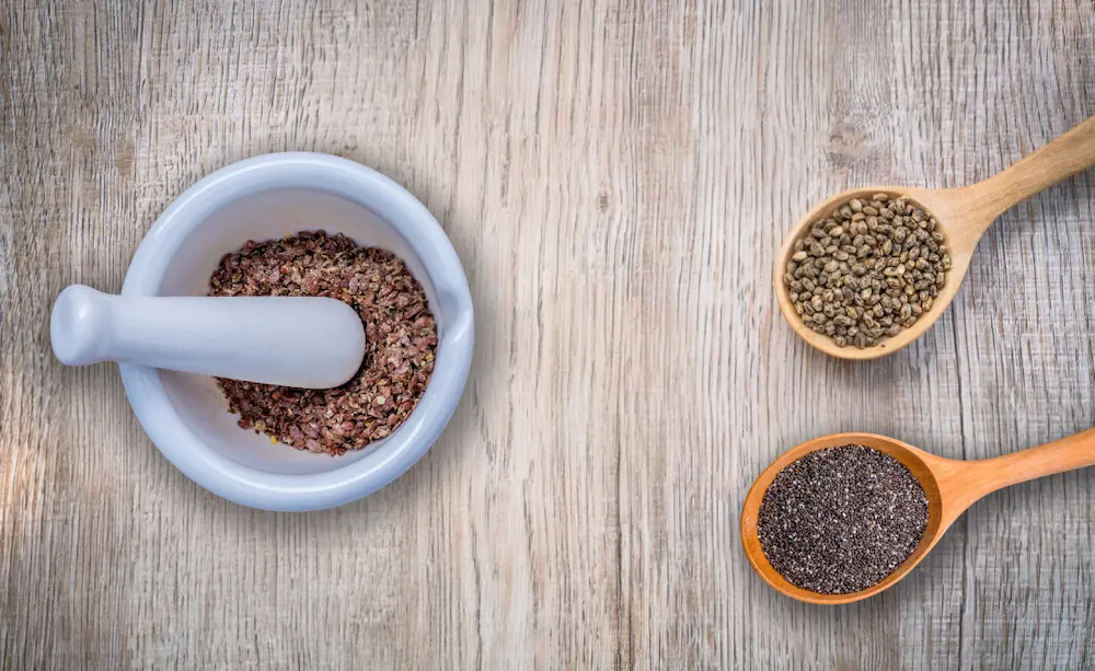 A white mortar and pestle crushing flaxseeds next to wooden spoons filled with raw hemp seeds and chia seeds, highlighting top plant-based omega-3 rich foods.