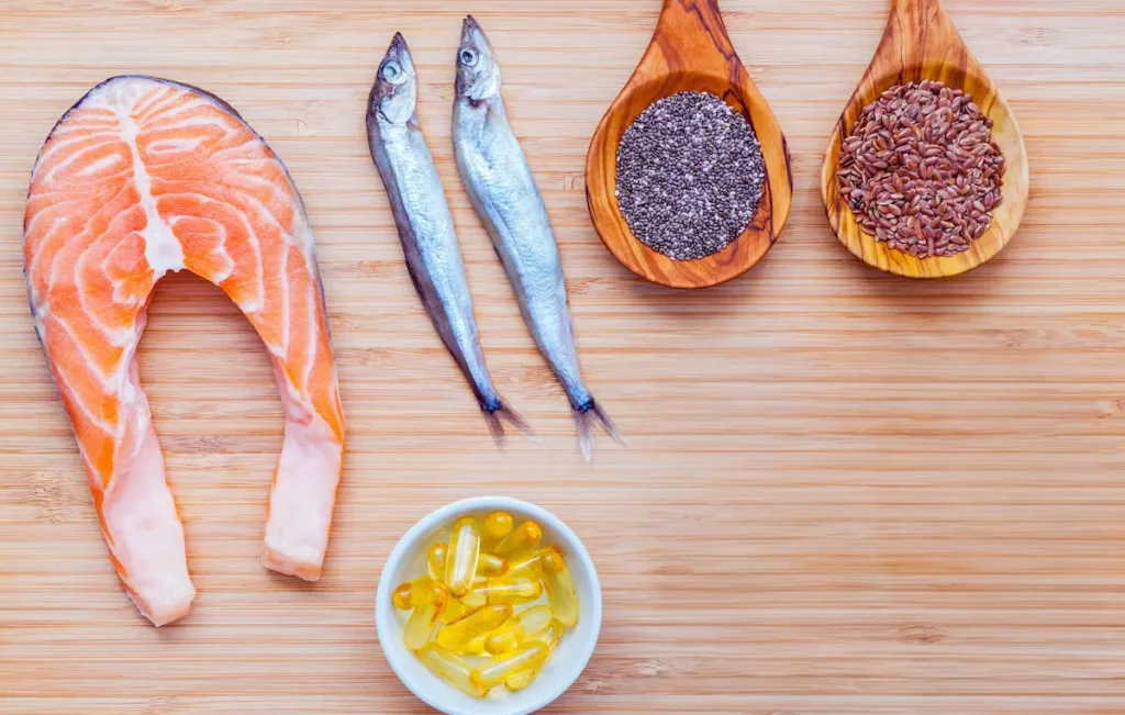 A wooden board displaying fresh salmon, small whole fish, chia seeds, flaxseeds, and supplement capsules representing the best omega-3 rich foods.