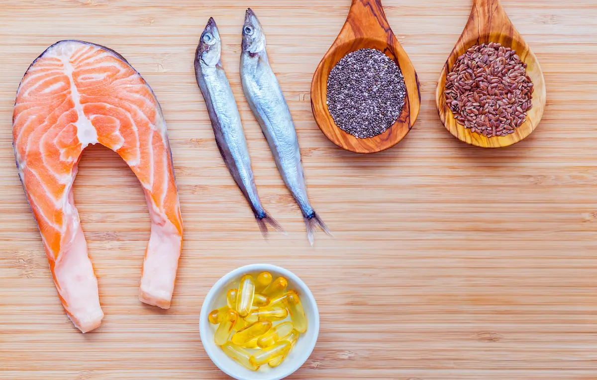 A wooden board displaying fresh salmon, small whole fish, chia seeds, flaxseeds, and supplement capsules representing the best omega-3 rich foods.