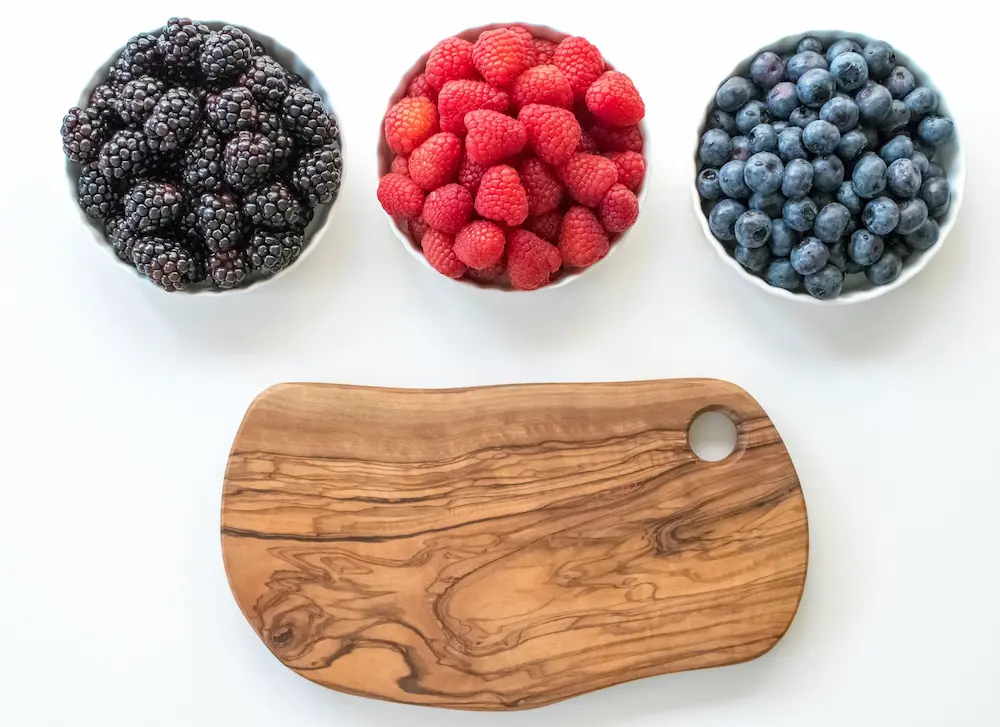 Three white bowls filled with fresh organic blueberries, raspberries, and blackberries arranged vertically next to a wooden cutting board.