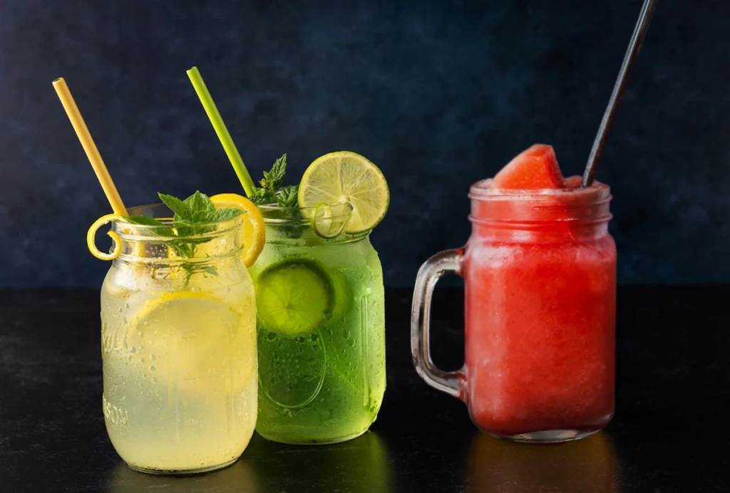 Three glass mason jars on a dark surface containing a yellow organic lemon and mint cooler, a green cucumber aloe vera drink, and a red watermelon basil juice.