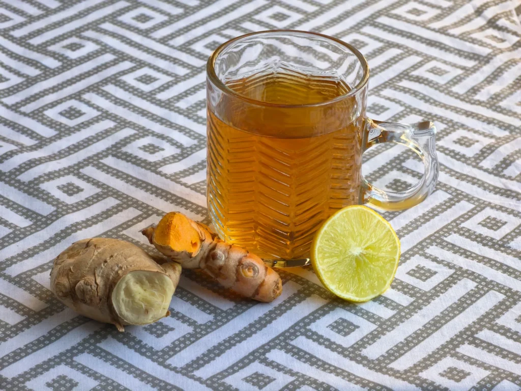 A glass mug of warm organic turmeric ginger tea resting on a geometric patterned tablecloth, surrounded by fresh ginger, sliced turmeric root, and a lemon wheel.