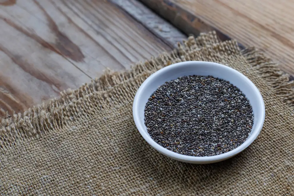 A small white bowl filled with raw, whole organic chia seeds resting on a rustic burlap cloth and wooden table.