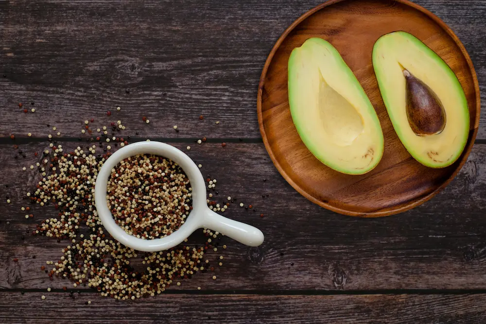 Top-down view of raw organic quinoa in a small white bowl next to a freshly halved avocado on a wood-patterned kitchen counter.