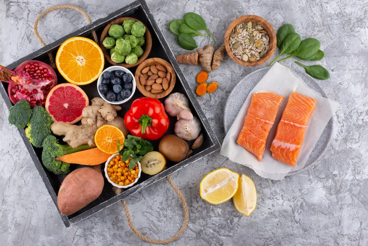 Colorful anti-inflammatory whole foods including raw salmon fillets, fresh berries, turmeric, ginger, and sweet potatoes arranged on and around a dark wooden tray.