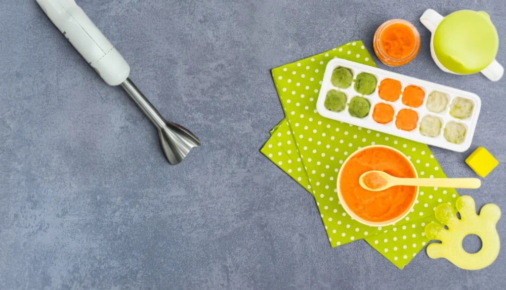 A top-down view of batch cooking homemade organic baby food, featuring a hand blender, a bowl of fresh orange carrot puree, and a silicone ice cube tray filled with frozen puree portions.