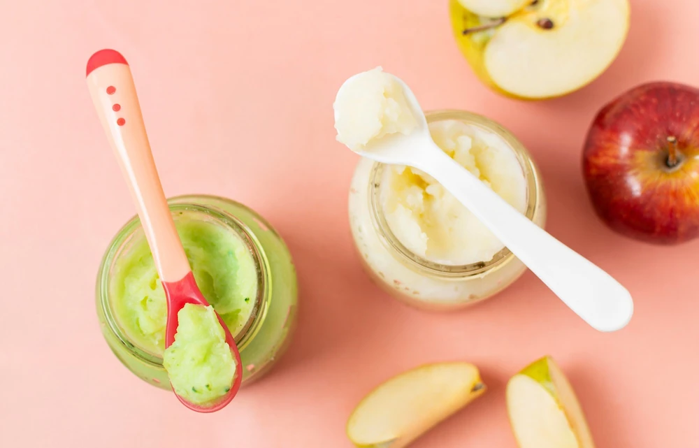 Fresh homemade avocado and apple baby food purees in glass jars with soft silicone spoons next to a fresh red apple and apple slices on a pink background.