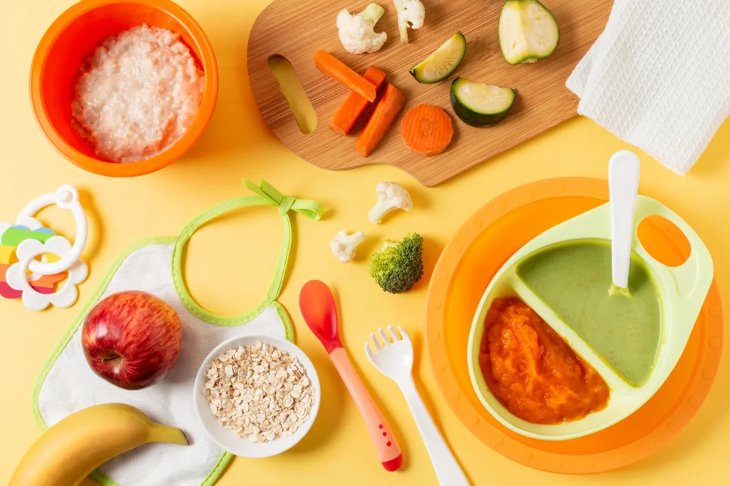 Homemade baby food in bowls with fresh fruits, vegetables, oats, and baby utensils on yellow background