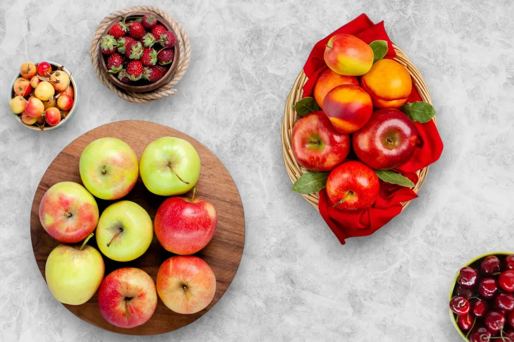 A top-down view of fresh strawberries, apples, peaches, and cherries in bowls on a light gray marble surface countertop to show fruits with high pesticide risk.
