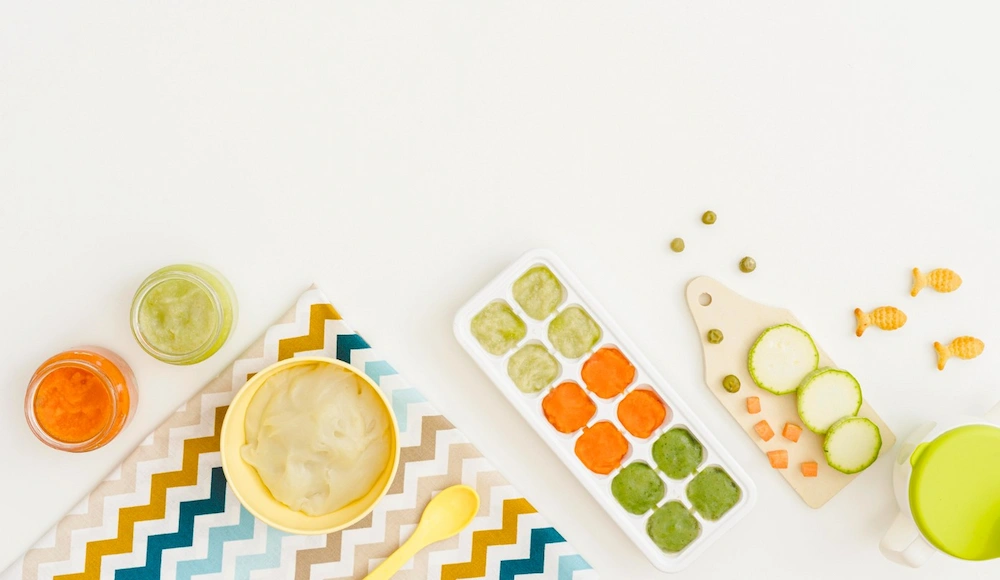 A clean preparation area showing safe baby food storage using small glass jars and silicone ice cube trays, alongside a small cutting board with fresh vegetables.