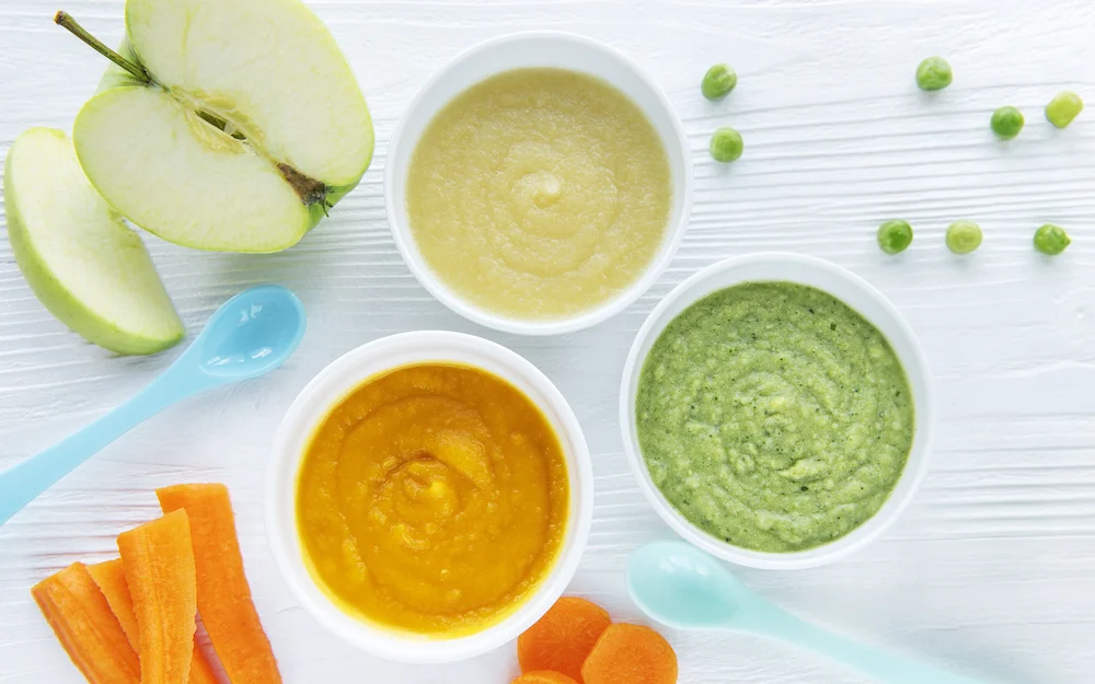 Three bowls of homemade stage two baby food purees in green, orange, and yellow, surrounded by fresh organic ingredients including sliced green apples, carrot sticks, and peas.