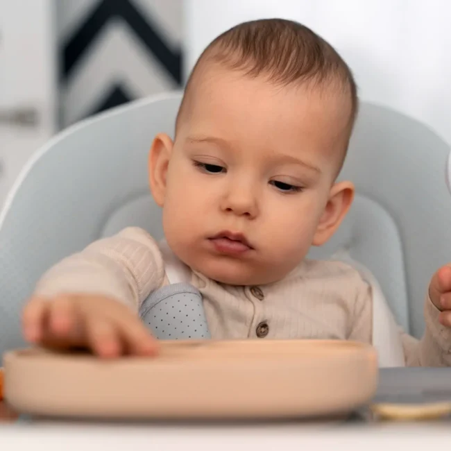 Baby sitting in high chair exploring first solid foods with spoon and plate