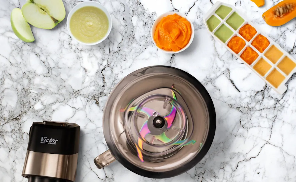 A top-down view of a Victor Electric blender, an ice cube tray filled with colorful vegetable purees, and fresh fruit on a marble counter to show how to batch cook baby food.
