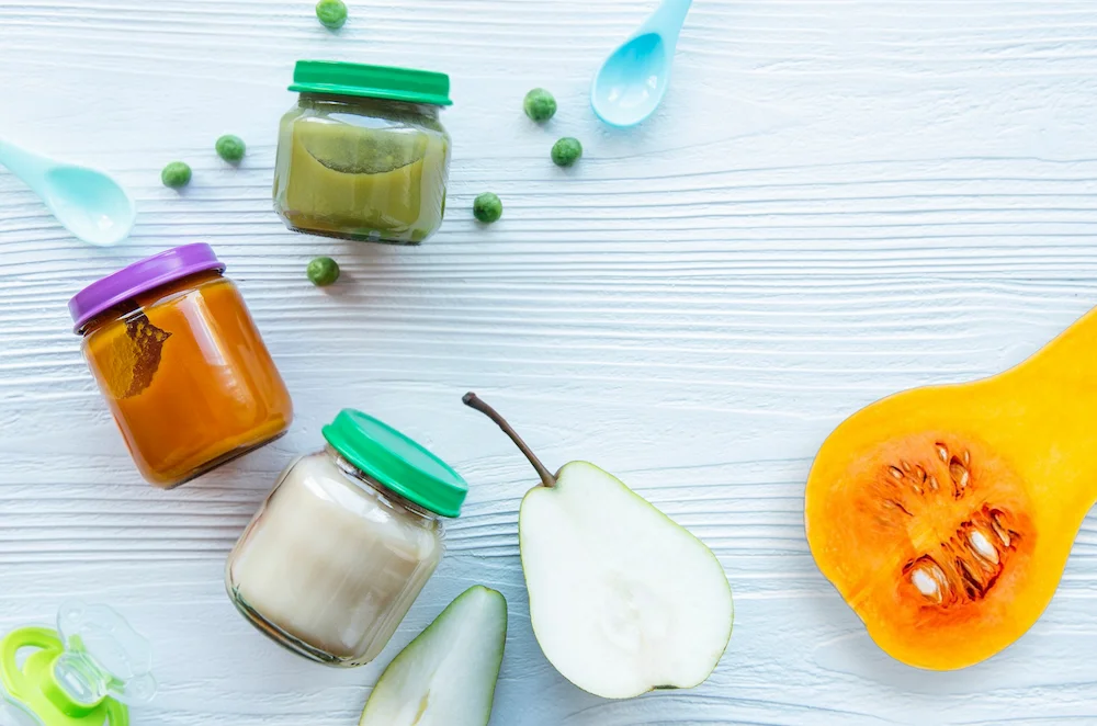Three glass jars of organic stage 1 baby foods featuring bright orange squash, yellow pear, and green pea purees next to fresh ingredients and baby spoons on a white wooden table.