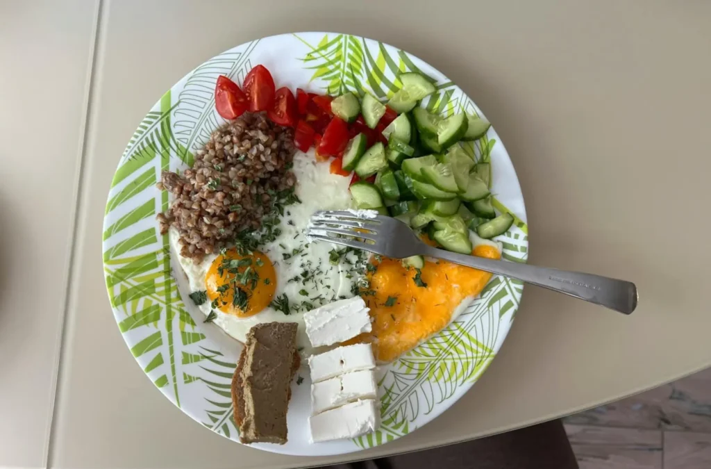 A healthy Mediterranean diet plate featuring fried eggs topped with herbs, whole grains, fresh cucumbers, cherry tomatoes, and sliced feta cheese.
