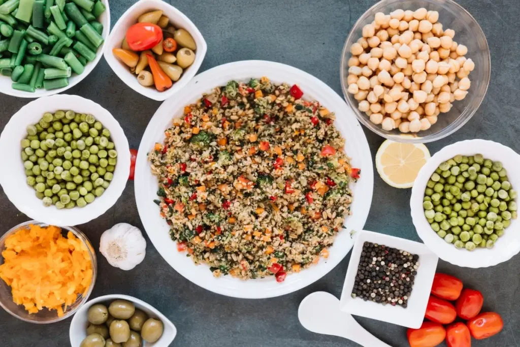 A top-down view of Mediterranean diet meal prep staples in bowls, featuring a whole grain salad surrounded by chickpeas, olives, fresh vegetables, lemon, and garlic.