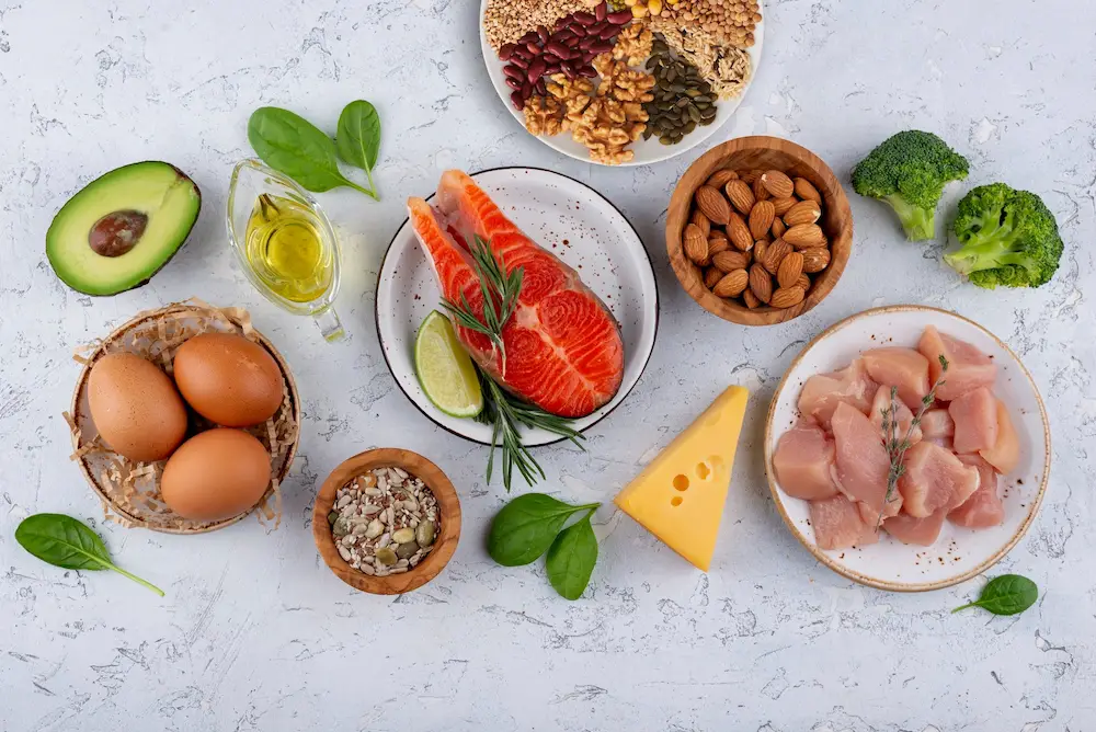 A top-down view of diabetic-friendly foods and pantry staples on a white textured surface, featuring high-quality proteins like fresh salmon and eggs, healthy fats like avocado and almonds, and low-glycemic carbohydrates including broccoli and mixed legumes.