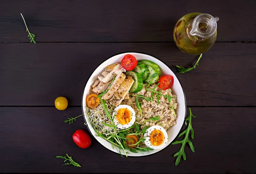 Top-down view of a balanced diabetic-friendly meal bowl with sliced chicken breast, boiled eggs, whole grains, cucumbers, and cherry tomatoes beside an olive oil cruet.