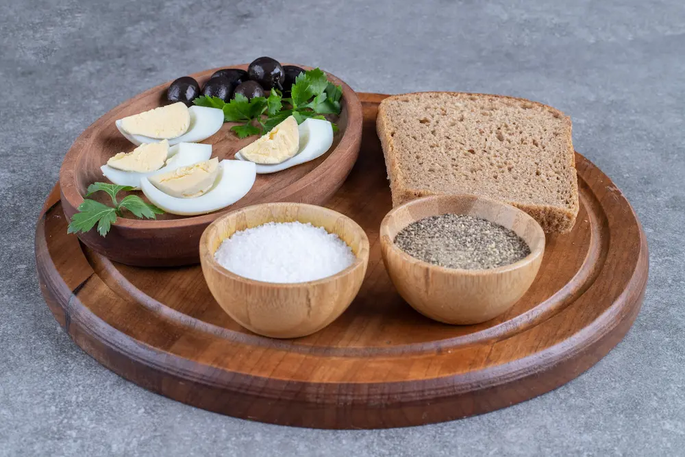 Sliced hard-boiled eggs served with black olives, fresh herbs, and a slice of whole-grain bread on a wooden serving board.