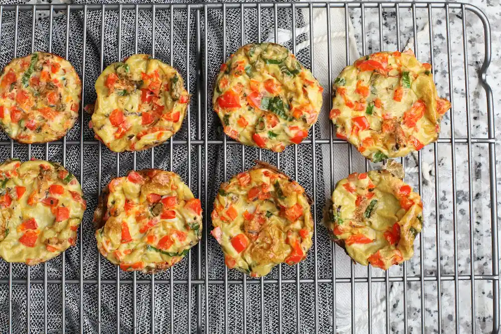 Eight thin baked high-protein egg bites with red bell peppers, spinach, and cheddar cheese cooling on a wire rack.
